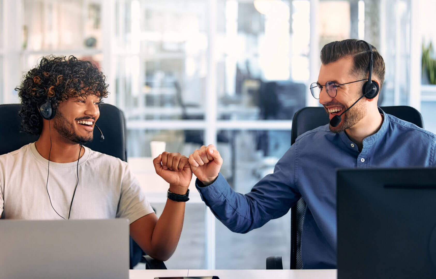 Two men wearing headsets sit at desks in an office, smiling and giving each other a fist bump.