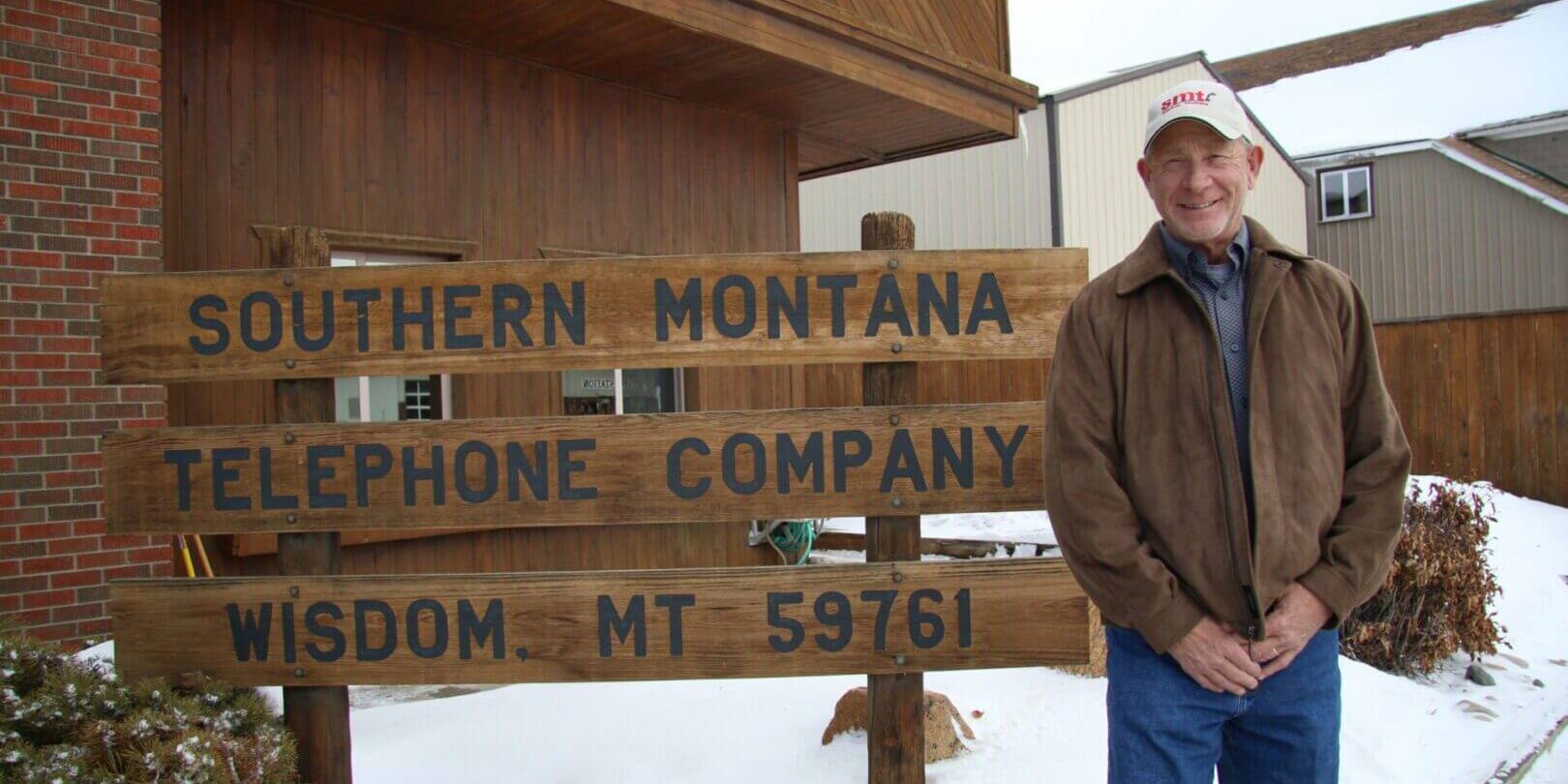 A man stands beside a wooden sign for Southern Montana Telephone Company in snowy weather.